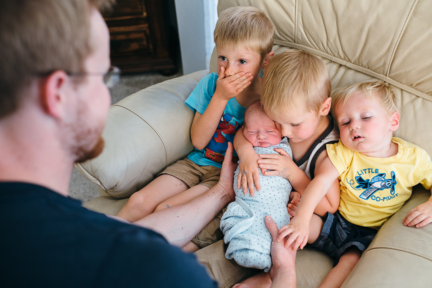 three young boys hold their new baby brother one looking shocked the other holding the baby tight and the third annoyed