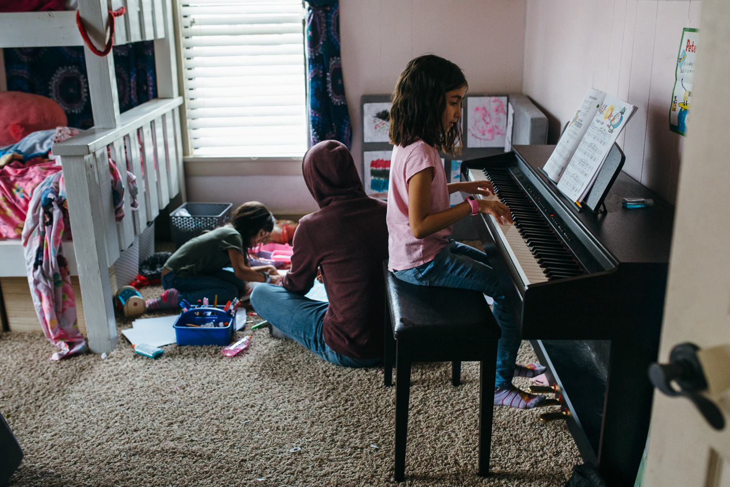a young girl plays the piano while another girl sits on the bedroom floor coloring