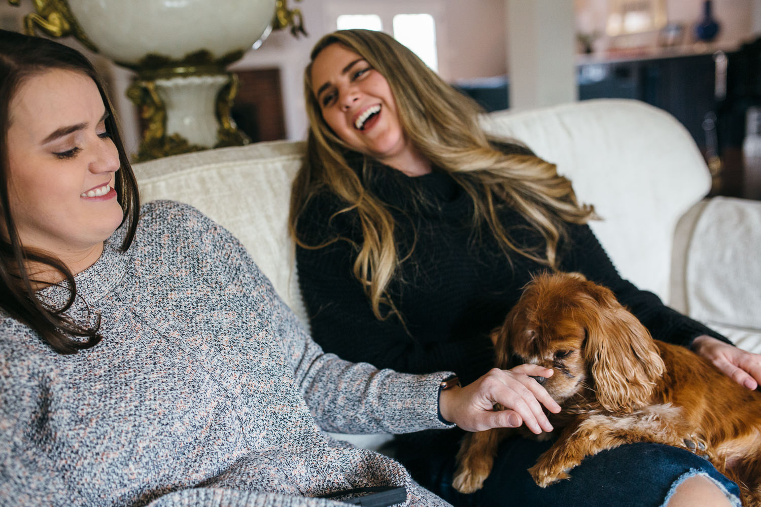 two women laughing on a cough while playing with a small dog on their lap